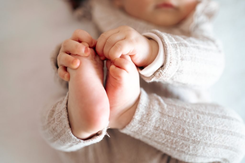 Le mani di una bimba piccola stringono i suoi piedini, sopra un maglioncino a coste color panna
