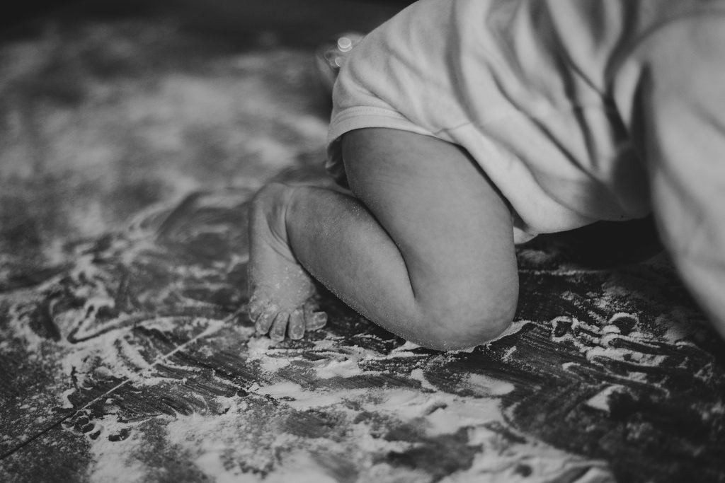 In bianco e nero, le ginocchia e i piedini nudi di una bimba che gattona sulla farina