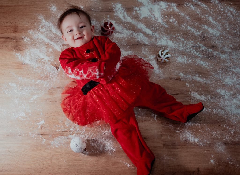Una bimba vestita di rosso con il tutù, sdraiata sul pavimento di legno coperto di farina, fra le palline di Natale