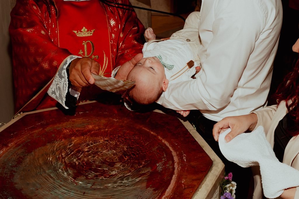 Il sacerdote in paramenti rossi versa l’acqua con una conchiglia sulla testa del bimbo, tenuto sopra il fonte battesimale