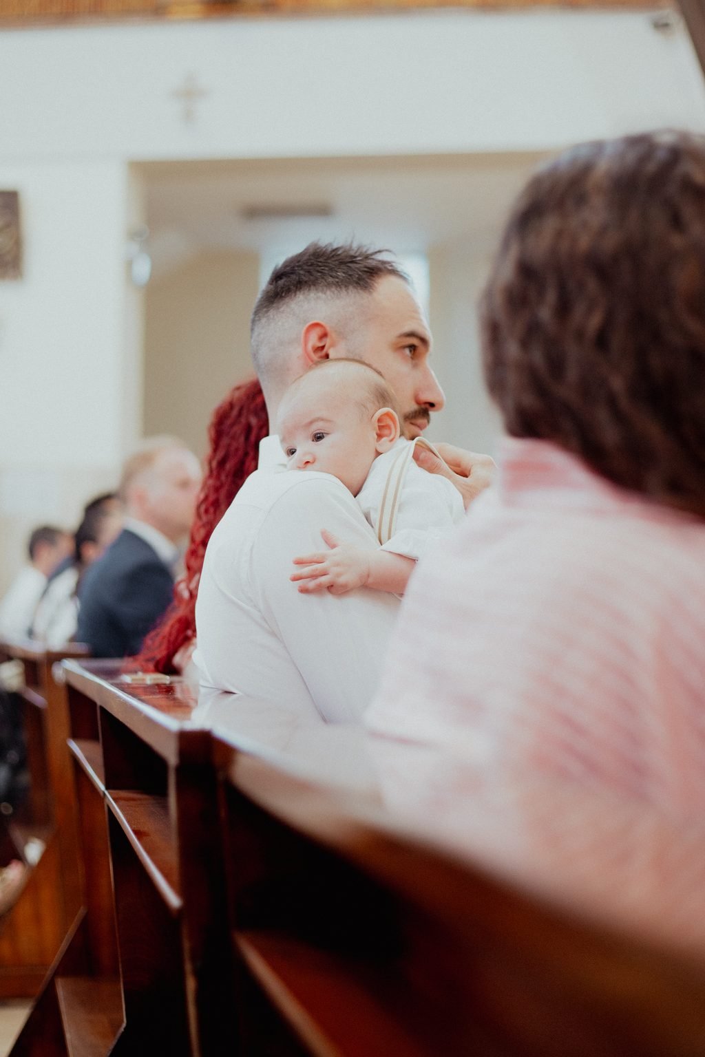 Il papà in camicia bianca tiene il bimbo sulla spalla, fra i parenti nei banchi della chiesa