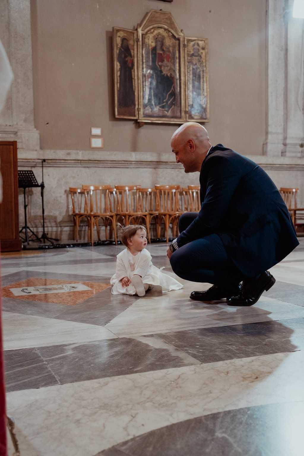 In una grande chiesa con il pavimento di marmo, il papà accovacciato tende le mani verso la bimba seduta per terra