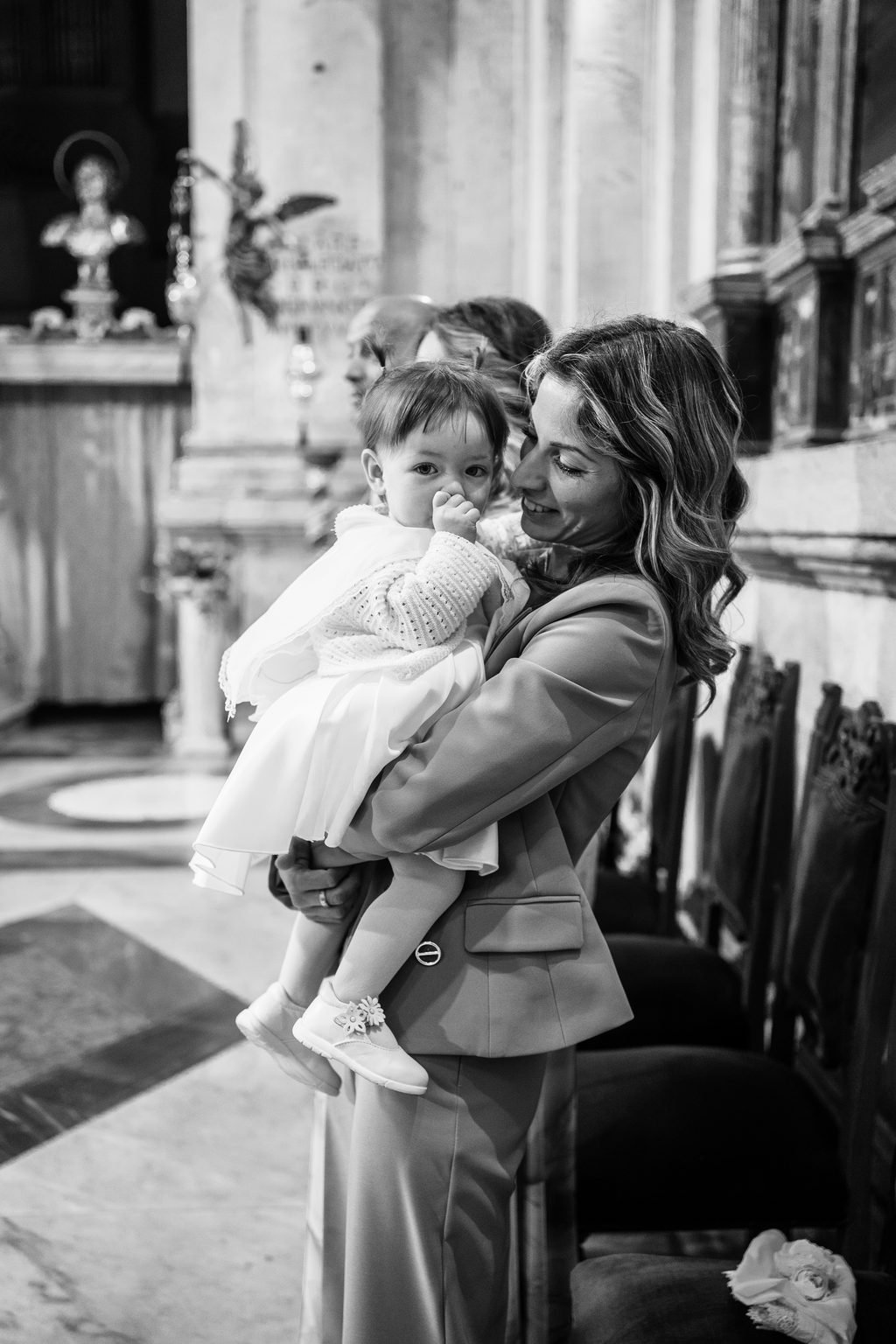 In bianco e nero, una donna stringe la bimba vestita di bianco fra le colonne della chiesa