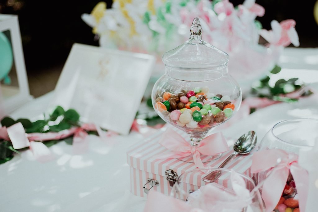 La confettata: un vaso di vetro pieno di confetti colorati sul tavolo bianco, fra fiori e nastri rosa