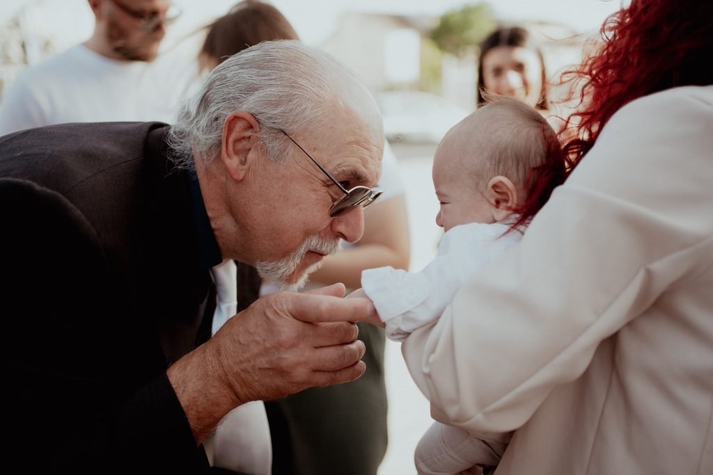 Un nonno con la barba bianca bacia la manina del nipotino tenuto in braccio dalla mamma