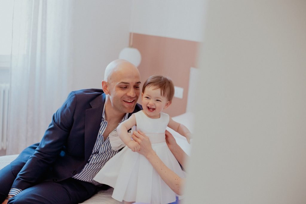 Il papà in abito blu, seduto sul letto, tiene in piedi la bimba vestita di bianco che ride