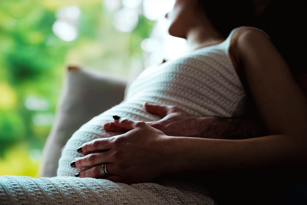 Due mani, una con la fede, appoggiate sulla pancia di una donna seduta vicino a una finestra con il verde fuori