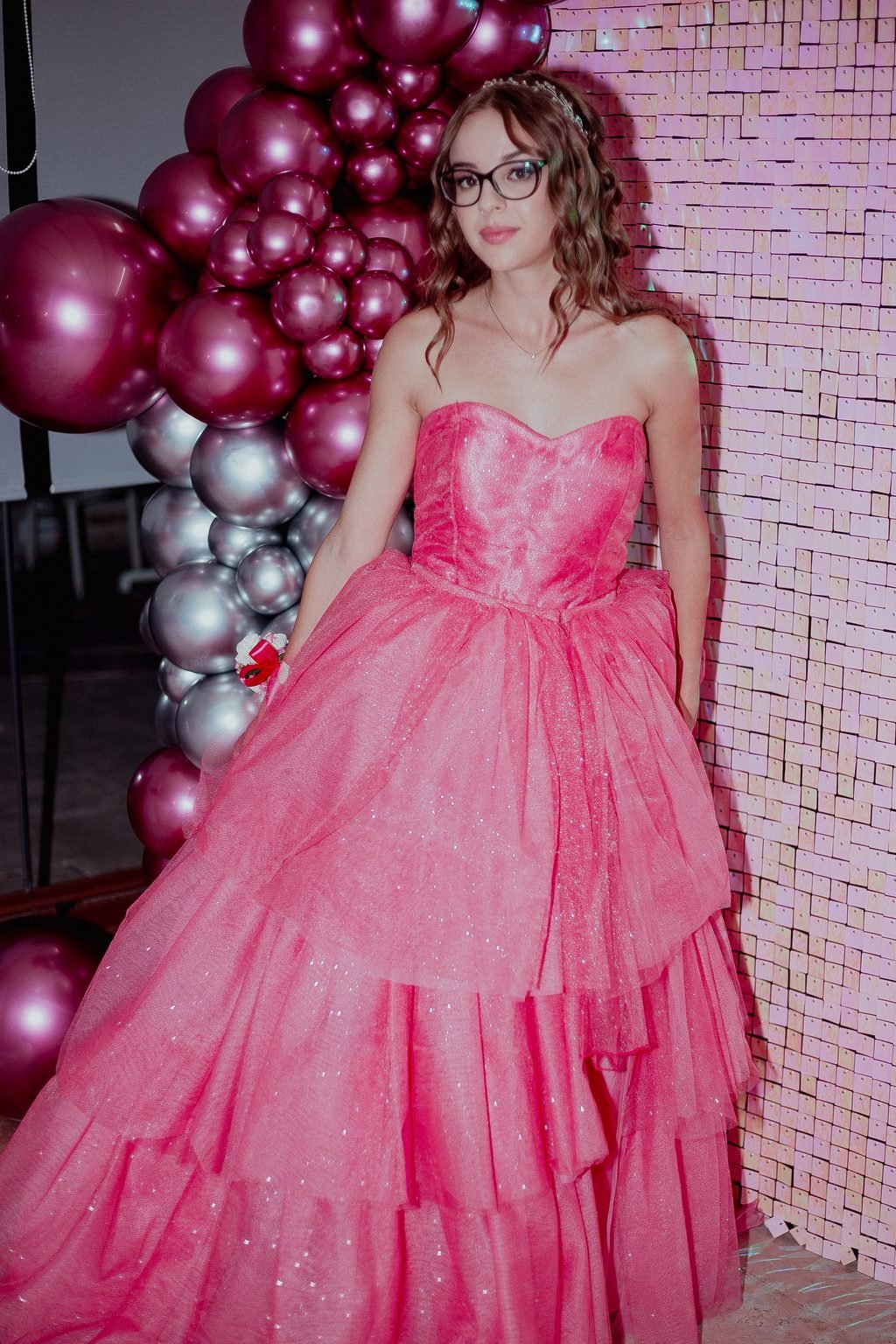 La festeggiata in abito rosa a balze posa davanti a una parete di palloncini rosa e argento