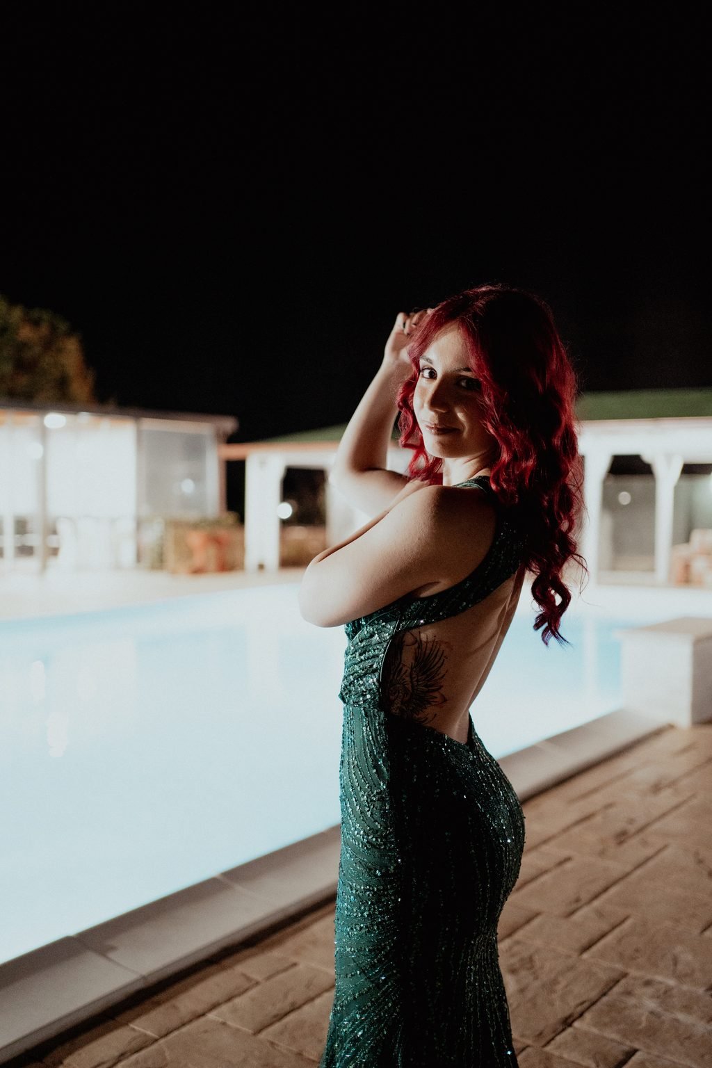 La festeggiata in abito verde, di spalle a bordo piscina, si gira verso l’obiettivo con una mano fra i capelli