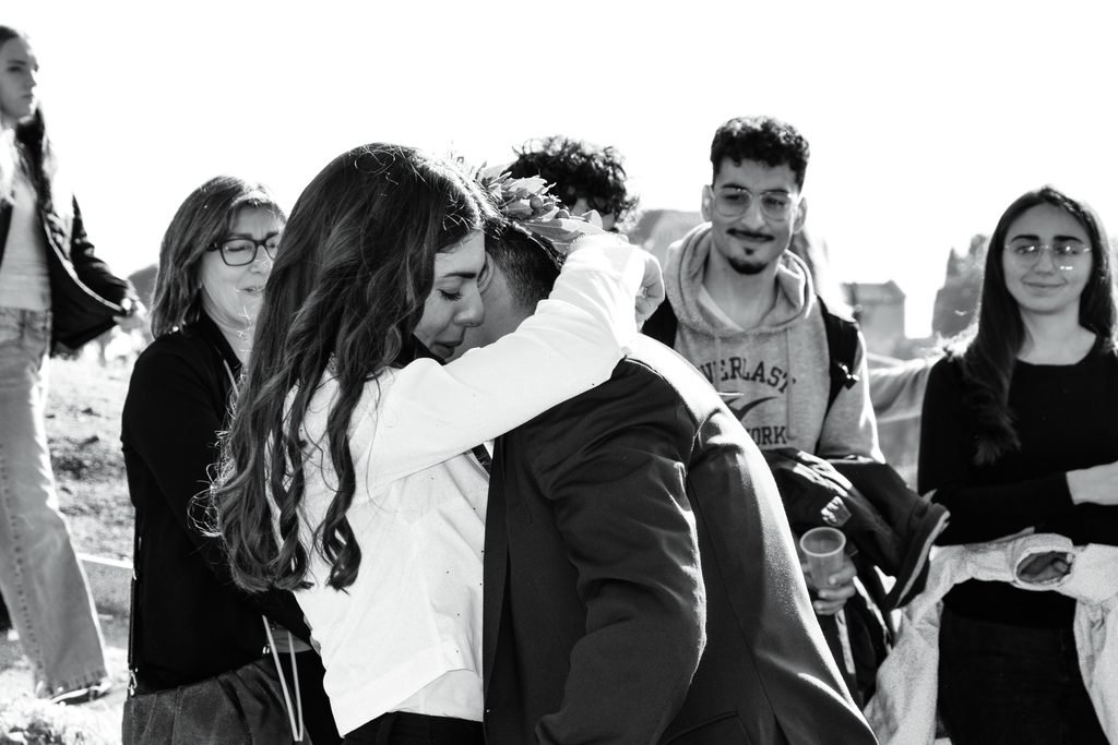 In bianco e nero, una ragazza abbraccia forte il neolaureato con la corona d’alloro, mentre gli amici intorno sorridono