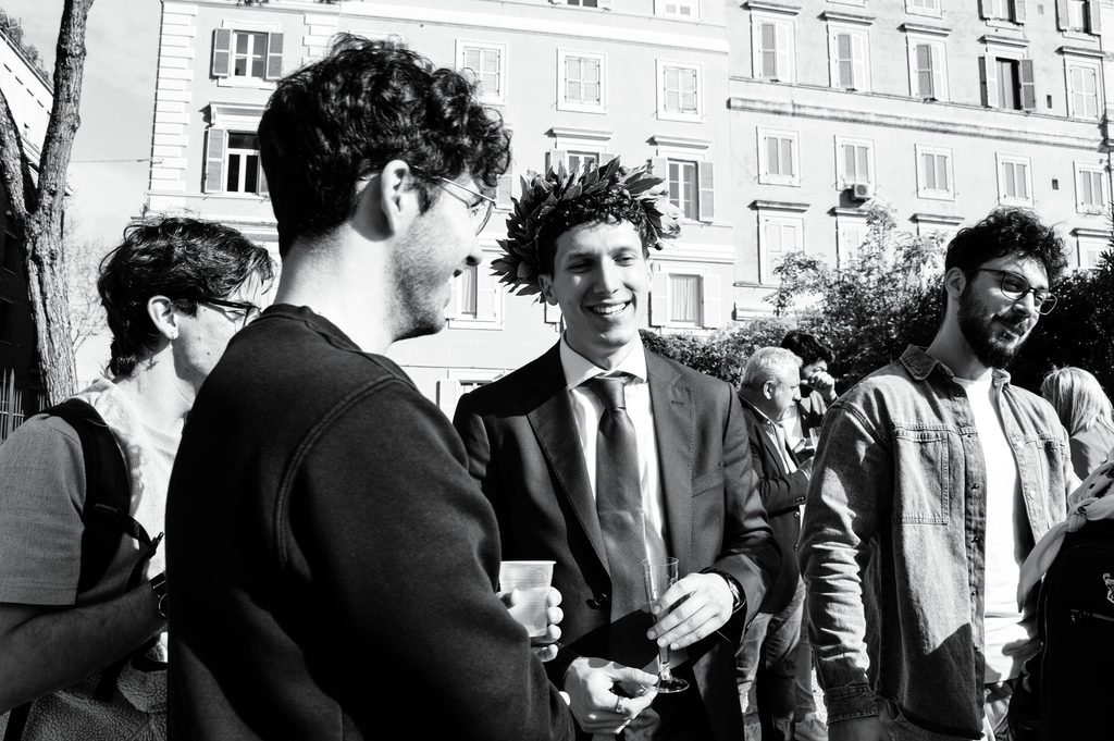 In bianco e nero, un neolaureato con la corona d’alloro sorride fra gli amici, con un bicchiere in mano, davanti ai palazzi di Roma