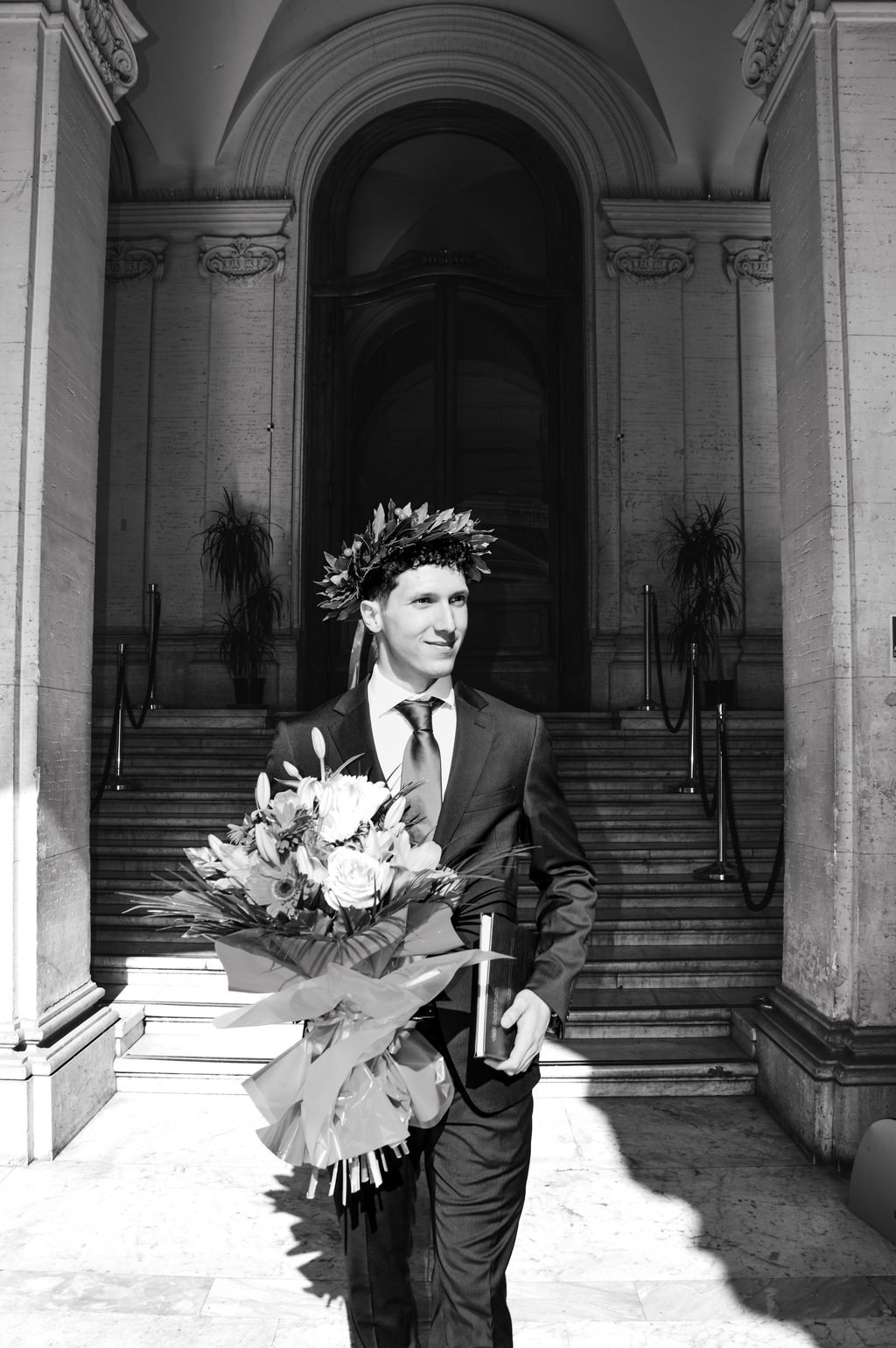 In bianco e nero, il neolaureato scende la scalinata dell’università con la corona d’alloro, i fiori e la tesi sotto il braccio
